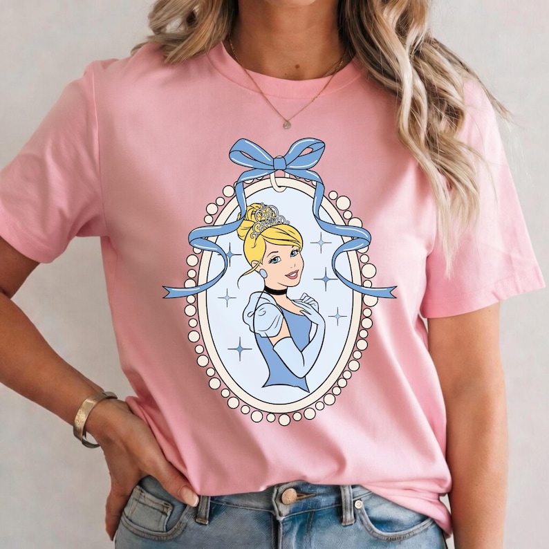K&ouml;nnte beinhalten: Hellrosa T-Shirt mit einem Cinderella-Motiv. Das Motiv ist ein ovales Bild mit hellblauem Hintergrund, einem Portr&auml;t von Cinderella und einer blauen Schleife. Die Prinzessin tr&auml;gt eine Tiara und ein blaues Kleid.