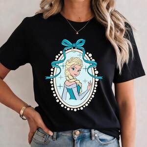 K&ouml;nnte beinhalten: Schwarzes T-Shirt mit einer Cartoon-Illustration einer blonden Frau in einem blauen Kleid und Zopf, umrahmt in einem Oval mit einem blauen Band. Das Design ist von wei&szlig;en Punkten und Kreuzen umgeben.