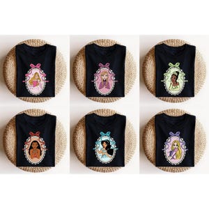 K&ouml;nnte beinhalten: Schwarze T-Shirts mit Prinzessinnenportr&auml;ts in ovalen Rahmen und Schleifen. Jedes Shirt zeigt eine andere Prinzessin, darunter Aurora, Rapunzel, Tiana, Pocahontas, Jasmin und Anna. Die Shirts werden auf gewebten Matten pr&auml;sentiert.