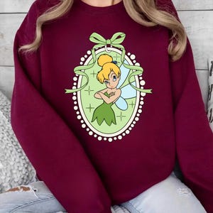 K&ouml;nnte beinhalten: Burgunderfarbenes Sweatshirt mit einem gr&uuml;n-wei&szlig;en ovalen Design mit einer Cartoon-Fee. Die Fee hat blondes Haar, ein gr&uuml;nes Kleid und blaue Fl&uuml;gel. Das Oval ist mit einem gr&uuml;nen Band und wei&szlig;en Punkten verziert.