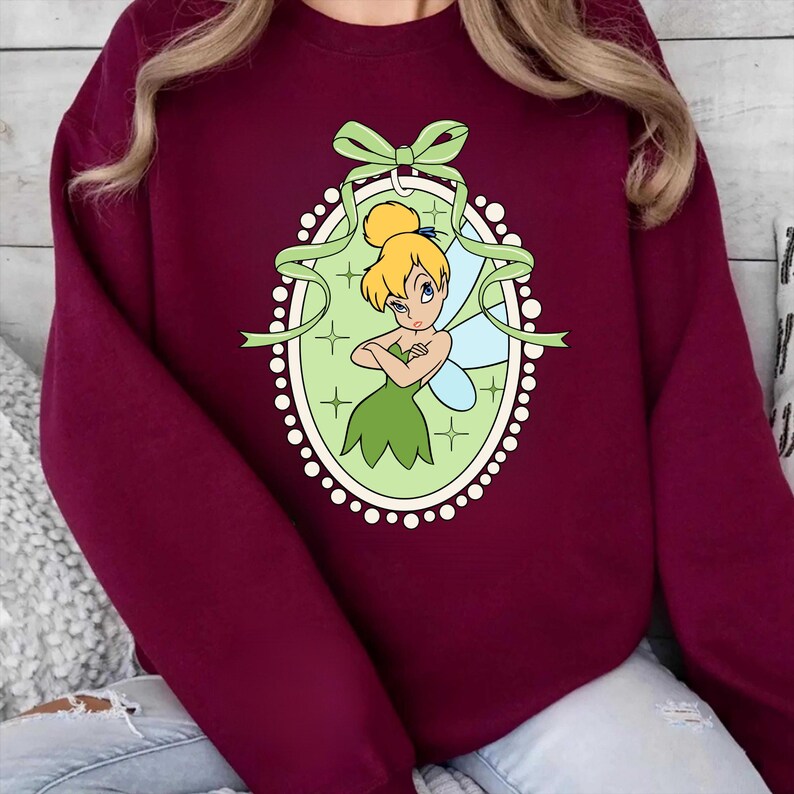 K&ouml;nnte beinhalten: Burgunderfarbenes Sweatshirt mit einem gr&uuml;n-wei&szlig;en ovalen Design mit einer Cartoon-Fee. Die Fee hat blondes Haar, ein gr&uuml;nes Kleid und blaue Fl&uuml;gel. Das Oval ist mit einem gr&uuml;nen Band und wei&szlig;en Punkten verziert.