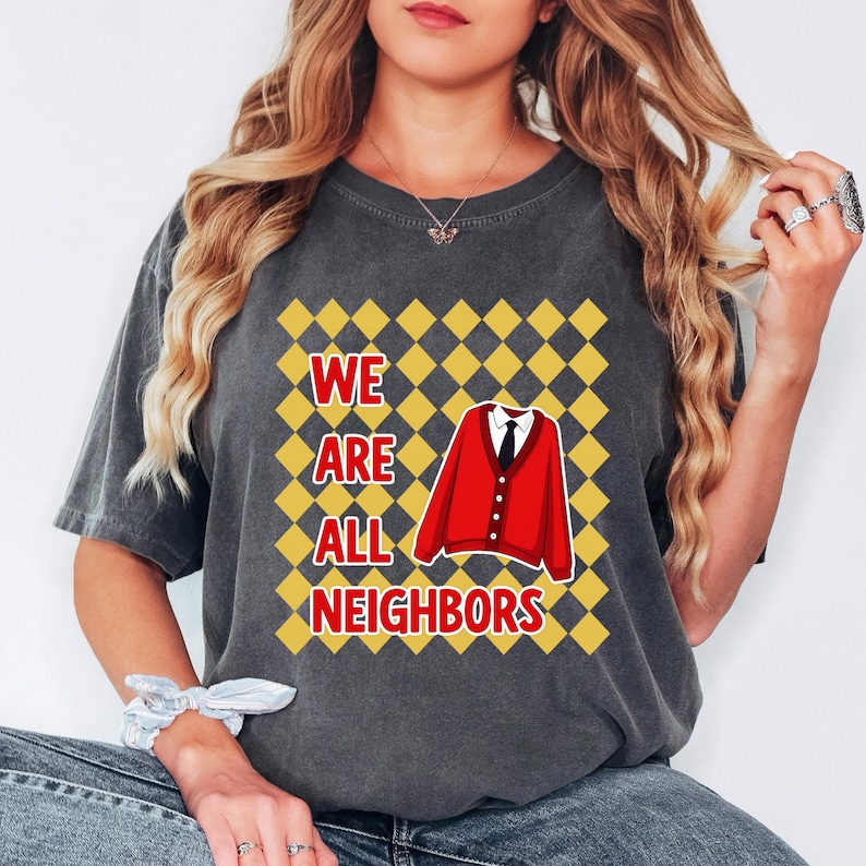 以下が含まれることがあります： 「WE ARE ALL NEIGHBORS」の白文字と赤いカーディガンが描かれた濃いグレーのTシャツ。グラフィックは黄と黒のチェック柄の背景に配置されています。カジュアルなスタイルのTシャツです。
