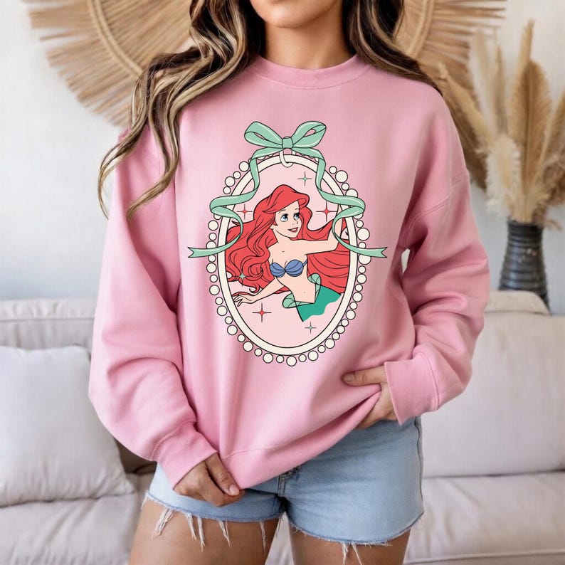 K&ouml;nnte beinhalten: Hellrosa Sweatshirt mit einer Cartoon-Illustration von Arielle, der Meerjungfrau. Die Grafik ist von einem hellrosa Oval mit einer mintgr&uuml;nen Schleife und einem Band umrahmt. Das lang&auml;rmlige Oberteil hat eine einfarbige rosa Farbe.