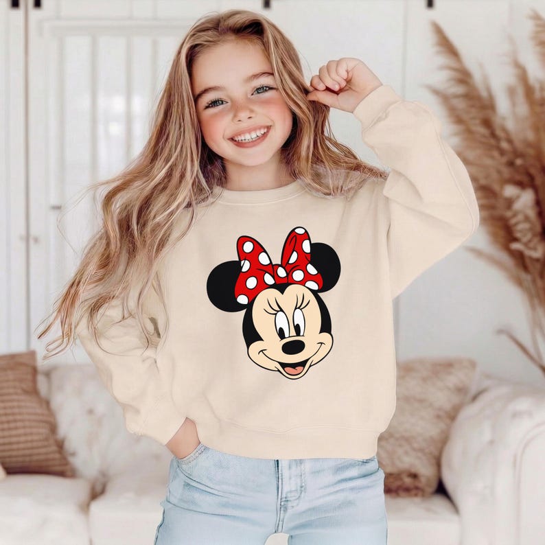 May include: A cream-coloured sweatshirt featuring a Minnie Mouse graphic. The graphic shows Minnie's face with a red bow with white polka dots. The sweatshirt has long sleeves and a crew neck.