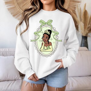 K&ouml;nnte beinhalten: Wei&szlig;es Sweatshirt mit einer Cartoon-Illustration einer Prinzessin in einem gr&uuml;nen und gelben ovalen Rahmen. Die Prinzessin hat dunkles Haar und tr&auml;gt eine Krone und Handschuhe. Ein gr&uuml;nes Band und eine Schleife befinden sich oben am Rahmen.