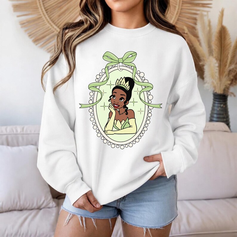 K&ouml;nnte beinhalten: Wei&szlig;es Sweatshirt mit einer Cartoon-Illustration einer Prinzessin in einem gr&uuml;nen und gelben ovalen Rahmen. Die Prinzessin hat dunkles Haar und tr&auml;gt eine Krone und Handschuhe. Ein gr&uuml;nes Band und eine Schleife befinden sich oben am Rahmen.