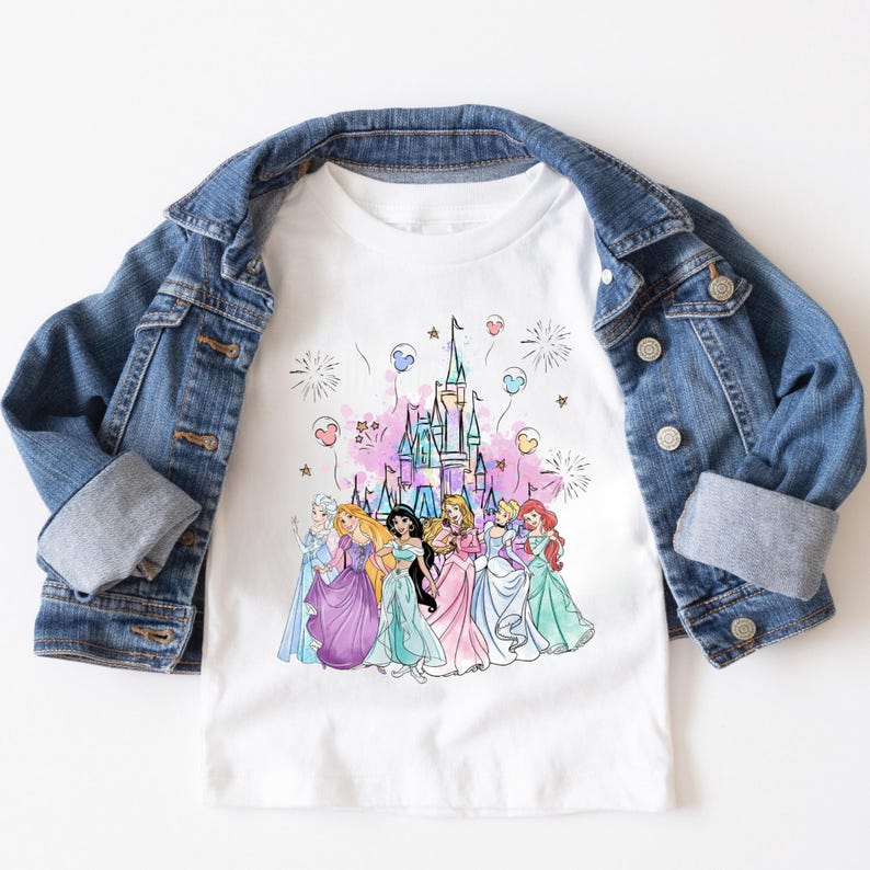 May include: White t-shirt featuring a colorful graphic of Disney princesses in front of a castle. The princesses are wearing gowns in various colors. A blue denim jacket is draped over the shirt.