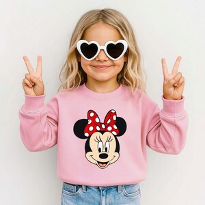 May include: A pink sweatshirt with a Minnie Mouse design, including a red and white polka dot bow. A child is wearing heart-shaped sunglasses and making a peace sign. The sweatshirt is a children's garment.