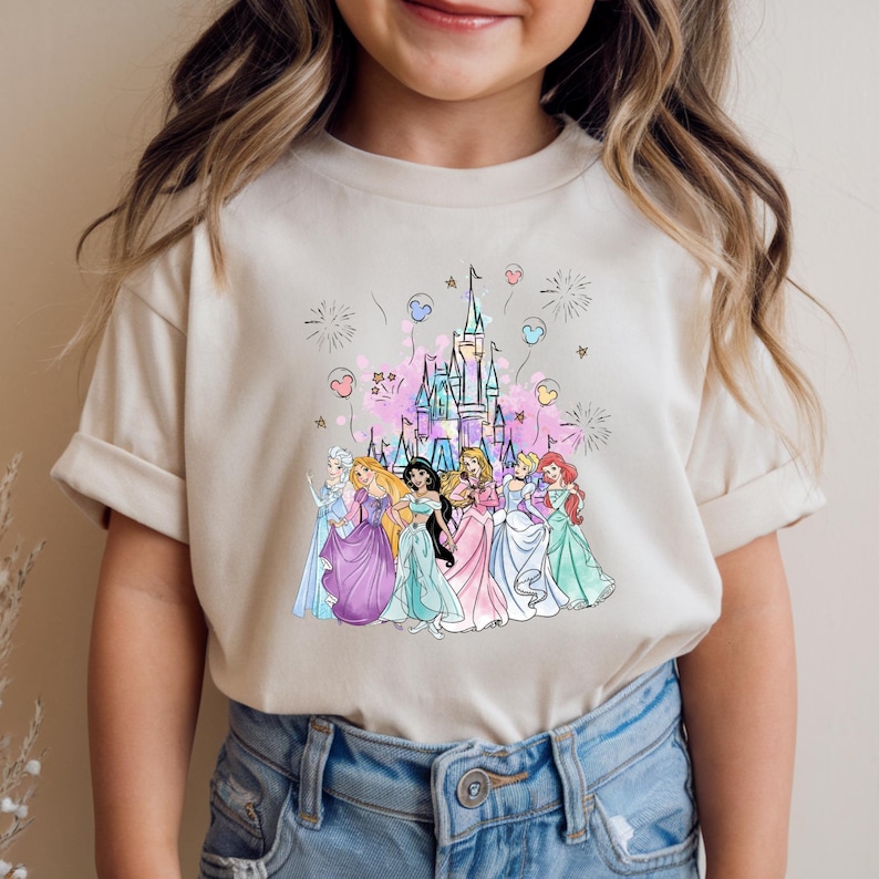 May include: A beige t-shirt featuring a colorful illustration of a castle and several princesses. The princesses are wearing gowns in various colors, and the castle is pink and purple. Fireworks and hearts are in the background.