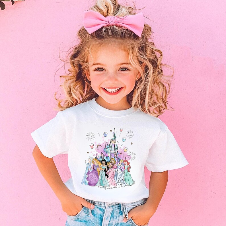 May include: A young girl wearing a white t-shirt featuring a colorful graphic of a castle and several princesses. She has blonde curly hair and a pink bow. The background is a solid pink.