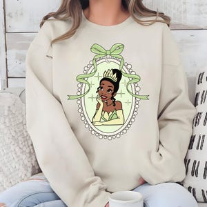 K&ouml;nnte beinhalten: Beiges Sweatshirt mit einer Cartoon-Illustration einer Prinzessin in einem gr&uuml;nen und goldenen Kleid, umrahmt von einem Oval mit gr&uuml;ner Schleife und Band. Die Prinzessin, mit einer Krone, st&uuml;tzt ihr Kinn auf ihre Hand. Das Design ist mittig auf dem Sweatshirt.