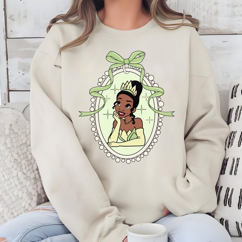 K&ouml;nnte beinhalten: Beiges Sweatshirt mit einer Cartoon-Illustration einer Prinzessin in einem gr&uuml;nen und goldenen Kleid, umrahmt von einem Oval mit gr&uuml;ner Schleife und Band. Die Prinzessin, mit einer Krone, st&uuml;tzt ihr Kinn auf ihre Hand. Das Design ist mittig auf dem Sweatshirt.