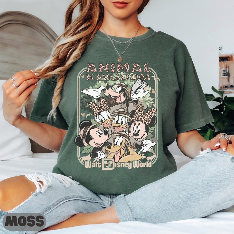 May include: Moss green t-shirt with a graphic of Disney characters, including Mickey and Minnie Mouse, Donald and Daisy Duck, and Goofy. The text "ANIMAL KINGDOM" and "Walt Disney World" are displayed on the front. The shirt is a moss green colour.