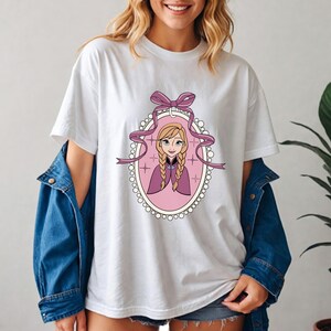 K&ouml;nnte beinhalten: Wei&szlig;es T-Shirt mit einer Cartoon-Illustration einer Figur mit blonden Z&ouml;pfen, die einen lila Umhang tr&auml;gt, in einem rosa ovalen Rahmen mit einem Band und einer Schleife. Das Design ist auf dem Shirt zentriert.