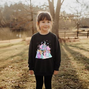 May include: Black sweatshirt featuring a colorful graphic of a castle and several princesses. The design includes pink, blue, and purple elements. The sweatshirt is worn by a young girl, and she is wearing black leggings.