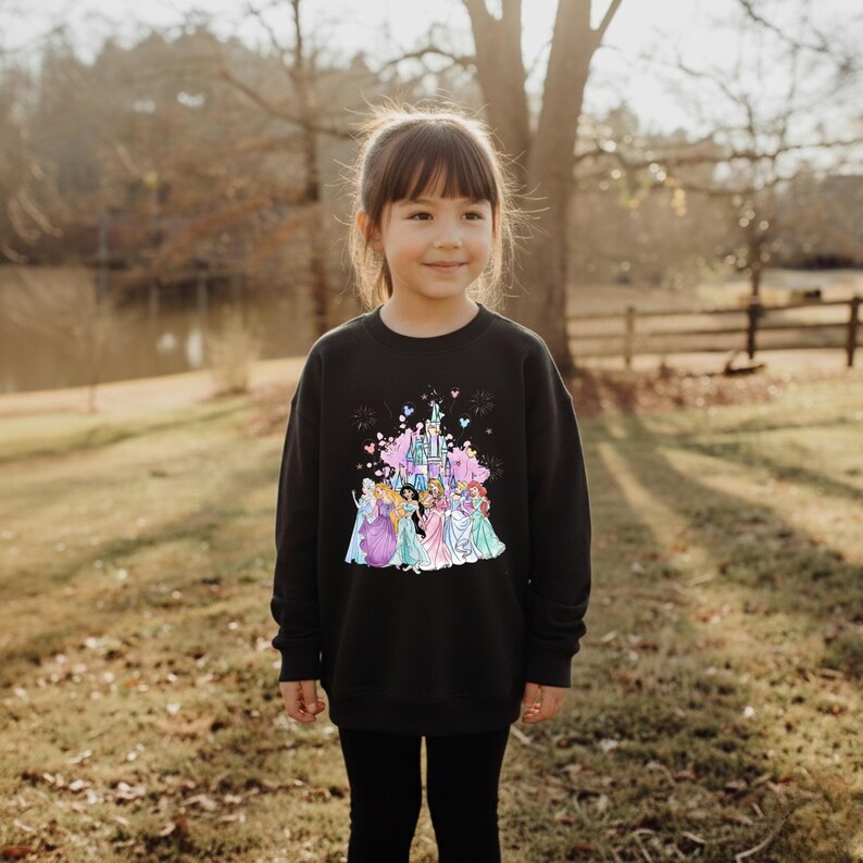 May include: Black sweatshirt featuring a colorful graphic of a castle and several princesses. The design includes pink, blue, and purple elements. The sweatshirt is worn by a young girl, and she is wearing black leggings.