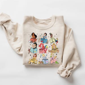 Peut inclure: Sweat-shirt cr&egrave;me avec un motif de princesses Disney lisant des livres. Chaque princesse est repr&eacute;sent&eacute;e avec ses compagnons animaux. Le motif comprend des &eacute;toiles et des titres de livres, cr&eacute;ant un th&egrave;me fantaisiste et litt&eacute;raire.