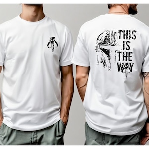 May include: A white short-sleeved t-shirt featuring a small black graphic of a helmet on the front left chest. The back of the shirt displays a large black graphic of a helmet with the text THIS IS THE WAY in a distressed, bold font.