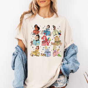 Peut inclure: T-shirt cr&egrave;me avec un motif color&eacute; de princesses Disney lisant des livres. Chaque princesse est repr&eacute;sent&eacute;e avec son propre livre, entour&eacute;e d'images associ&eacute;es. Le t-shirt a des manches courtes et une coupe d&eacute;contract&eacute;e.