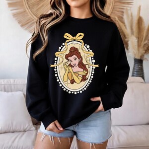 K&ouml;nnte beinhalten: Schwarzes Sweatshirt mit einer Cartoon-Illustration einer Prinzessin in einem gelben Kleid und Handschuhen, umrahmt von einem gelben Band und einem ovalen Rand mit wei&szlig;en Punkten. Die Prinzessin hat braune Haare und l&auml;chelt.