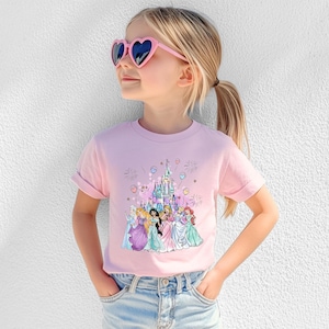 May include: A light pink t-shirt featuring a colorful graphic of a castle and several princesses. The shirt is worn by a child wearing pink heart-shaped sunglasses and blue jeans.