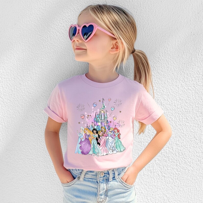 May include: A light pink t-shirt featuring a colorful graphic of a castle and several princesses. The shirt is worn by a child wearing pink heart-shaped sunglasses and blue jeans.
