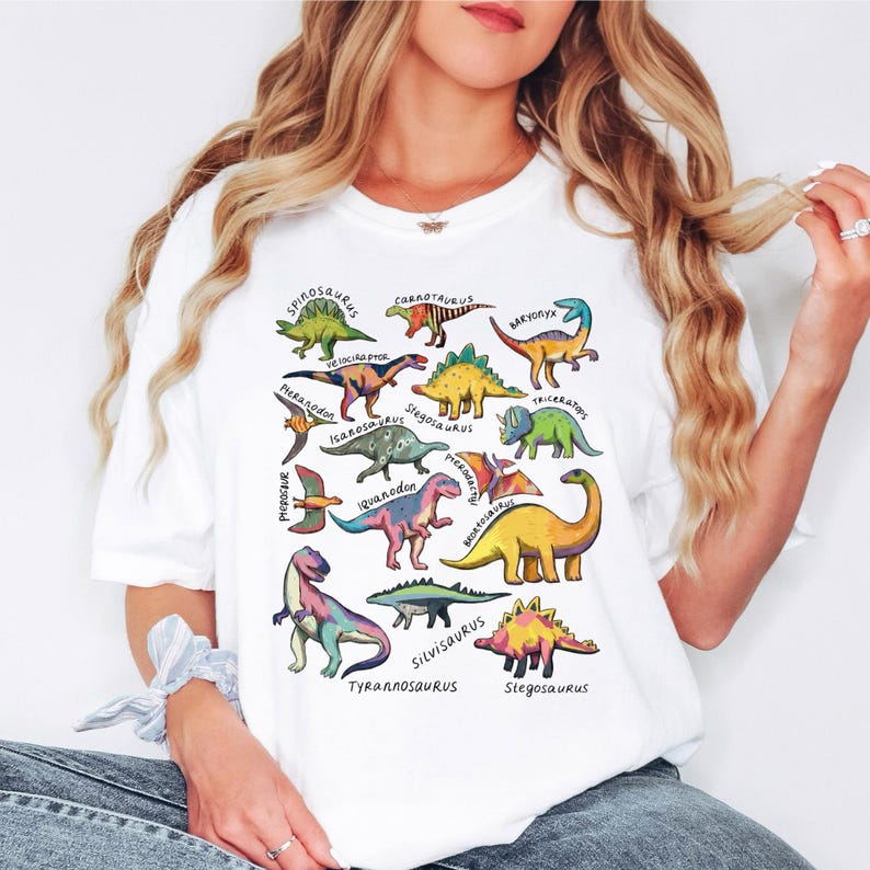 May include: White t-shirt featuring colorful illustrations of various dinosaurs with their names printed below. The dinosaurs are in different colors and poses, including a green Spinosaurus and a yellow Stegosaurus.