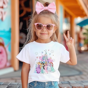 May include: A young child wearing a white t-shirt featuring a colorful graphic of Disney princesses in front of a castle. The child is also wearing pink sunglasses and a pink bow. The child is giving a peace sign.