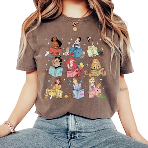 Peut inclure: T-shirt marron avec un motif color&eacute; de princesses Disney lisant des livres. Chaque princesse est repr&eacute;sent&eacute;e avec son nom sur le livre, entour&eacute;e d'images associ&eacute;es. Le t-shirt est de style d&eacute;contract&eacute; et confortable.