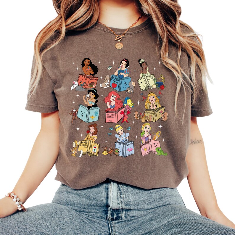 Peut inclure: T-shirt marron avec un motif color&eacute; de princesses Disney lisant des livres. Chaque princesse est repr&eacute;sent&eacute;e avec son nom sur le livre, entour&eacute;e d'images associ&eacute;es. Le t-shirt est de style d&eacute;contract&eacute; et confortable.