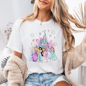 May include: White t-shirt featuring a colorful illustration of Disney princesses in front of a castle. The design includes fireworks, balloons, and a pink and purple color scheme. The princesses are wearing gowns in various colors.