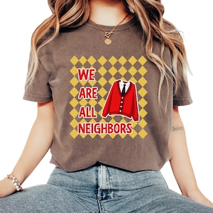 以下が含まれることがあります： 「WE ARE ALL NEIGHBORS」の文字が白と黄色でプリントされた茶色のTシャツ。赤いカーディガンと黒いネクタイがデザインの中央にあり、黄とオレンジのひし形模様が背景になっています。