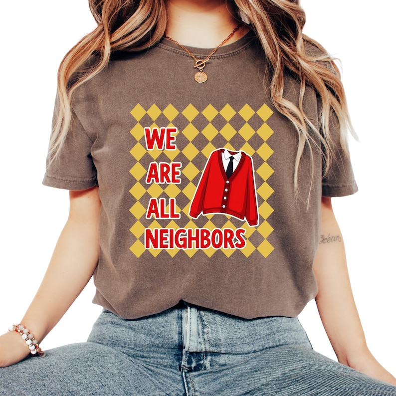 以下が含まれることがあります： 「WE ARE ALL NEIGHBORS」の文字が白と黄色でプリントされた茶色のTシャツ。赤いカーディガンと黒いネクタイがデザインの中央にあり、黄とオレンジのひし形模様が背景になっています。