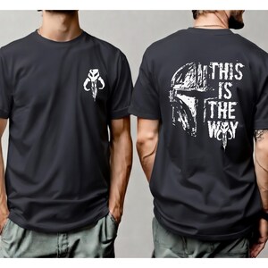 May include: A dark gray short-sleeved t-shirt featuring a white graphic of a helmeted skull on the left chest. The back of the shirt displays a large white graphic of a helmet and the text THIS IS THE WAY in a distressed, bold font style.