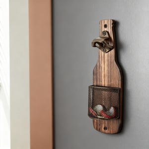 Puede incluir: Un abridor de botellas de pared con forma de botella de madera con una cesta de malla metálica para tapones. El abridor es de madera oscura y tiene un abridor de color bronce adjunto en la parte superior. La cesta contiene varias tapas.