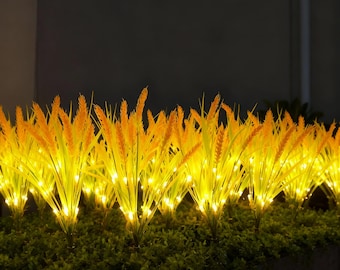 May include: Illuminated outdoor wheat grass. The artificial plants have yellow-orange wheat stalks and green leaves. Small, warm white LED lights are interspersed throughout the wheat grass, creating a soft glow. The background is dark.