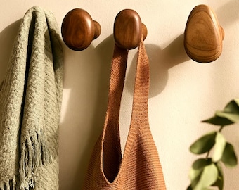 May include: Three wooden wall hooks in varying shapes and sizes are mounted on a light-colored wall. A woven brown tote bag hangs from one hook, and a green fringed blanket is draped near another. The hooks are made of a warm-toned wood.