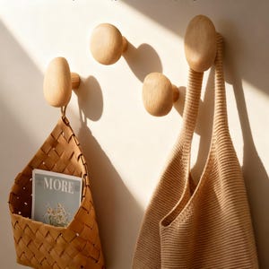 Puede incluir: Tres perchas de pared de madera con acabado natural están montadas en una pared de color claro. Una cesta tejida y un bolso tote beige cuelgan de las perchas. La cesta contiene una revista con la palabra "MORE" visible.