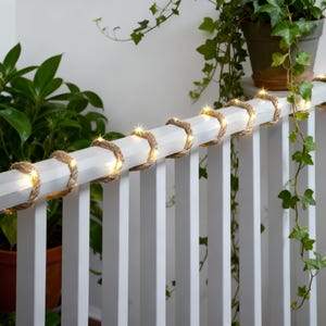 May include: A white wooden railing adorned with a natural rope and string lights. The rope is wrapped around the railing, and the lights are evenly spaced. Green plants in pots are in the background, creating a natural, decorative theme.