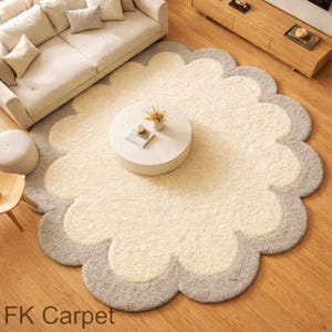 Può includere: Un grande tappeto a forma di fiore con un centro color crema e un bordo grigio. Il tappeto è posizionato su un pavimento in legno ed è circondato da mobili, tra cui un divano e un tavolino. Il testo "FK Carpet" è visibile in basso a sinistra.