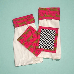 May include: A set of white kitchen towels and pot holders with a vibrant pink and gold leopard print design. One pot holder features a black and white checkered pattern. The set is designed for kitchen use.
