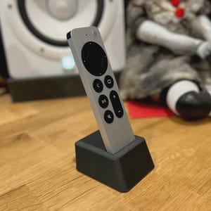 May include: A silver remote control with black buttons and a circular control pad is displayed in a black stand. The remote is angled slightly towards the viewer. The stand is on a wooden surface.