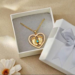 May include: A gold-tone necklace featuring a heart-shaped pendant lined with small clear stones. Inside the heart are two baby foot charms, one engraved with the name Courtravin and the other with Derriqua, centered by a small blue stone.