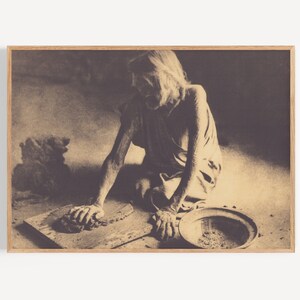 May include: A black and white photograph of a person sitting on the floor and working with clay. The person is using their hands to shape the clay on a wooden board. There is a bowl of clay in front of them.
