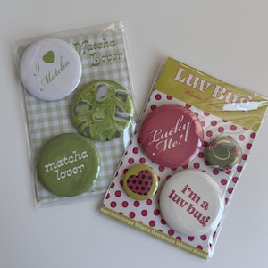 May include: Two sets of circular pin-back buttons in clear packaging. One set features green and white designs with text reading I Matcha, Matcha Lover, and a green heart. The other set features red and white designs with text reading Luv Bug, Lucky Me, and i'm a luv bug.