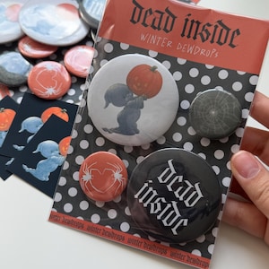 May include: A set of four round pinback buttons on a black and white polka dot backing card labeled dead inside winter dewdrops. The buttons feature a pumpkin-headed figure, a spider web, a heart with spider legs, and the text dead inside.