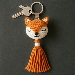 Puede incluir: Un llavero de zorro hecho a mano, naranja y blanco, con un llavero y llaves plateadas. El zorro tiene los ojos cerrados y una nariz negra. Una larga borla naranja cuelga debajo de la cabeza del zorro. El llavero está hecho de hilo.