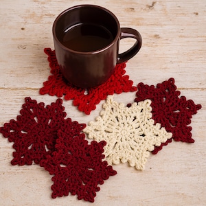 May include: A brown mug filled with a dark liquid sits on a red crocheted snowflake coaster. Several other snowflake coasters in red and cream colors are arranged nearby on a light wood surface. The coasters are handmade.