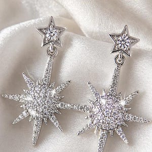 May include: A pair of silver-toned starburst earrings. Each earring features a small star at the top and a larger starburst design below, both encrusted with sparkling clear crystals. The earrings dangle from a small loop.