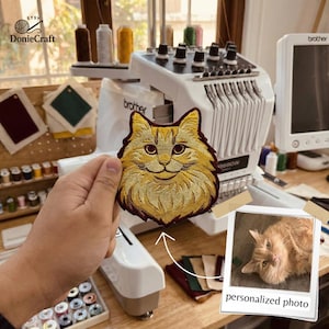 May include: Embroidered patch featuring a smiling cat face in shades of gold and brown. The patch is held in front of an embroidery machine. A small photo with the text "personalized photo" is also visible.